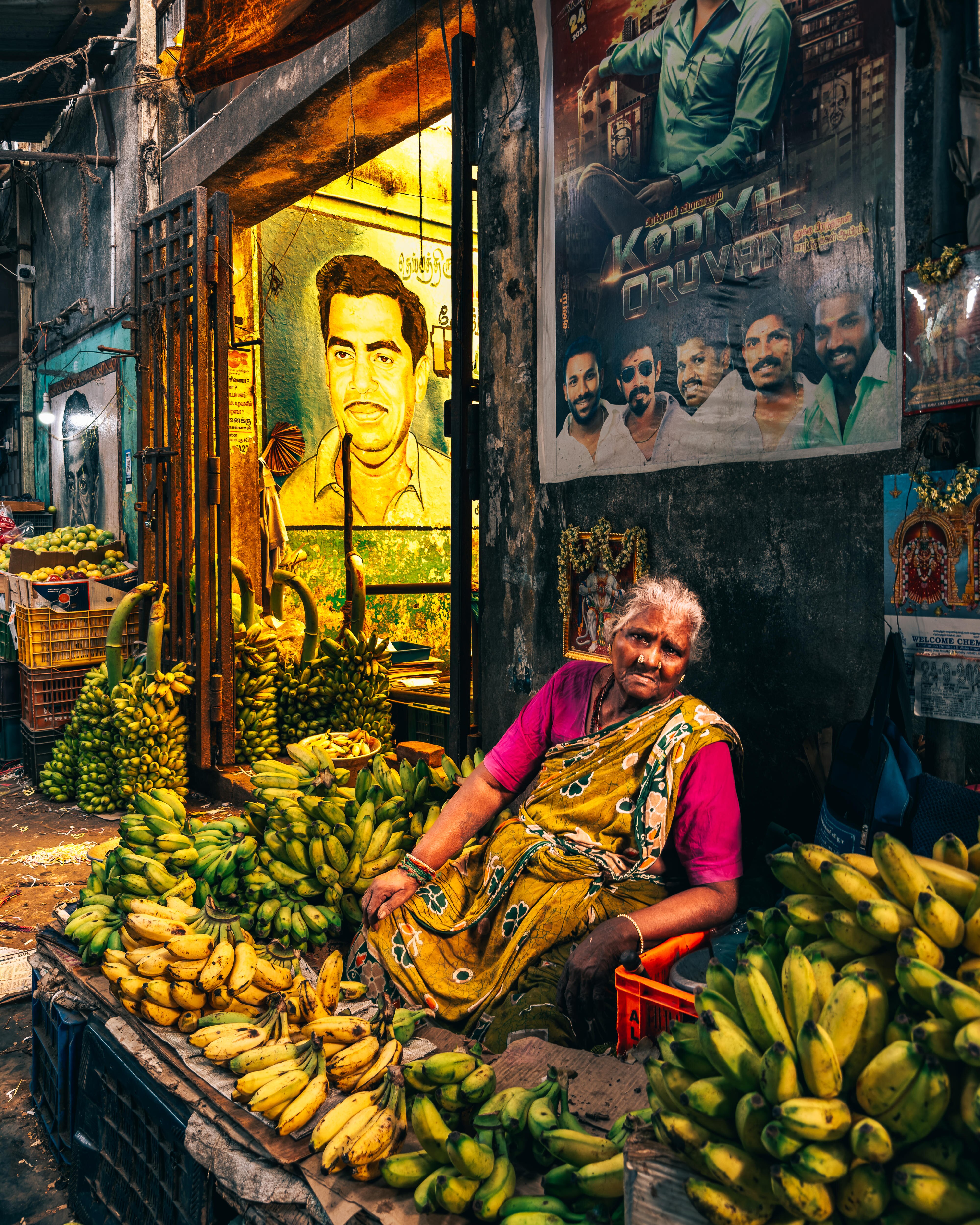 A woman wearing a yellow sari sits among bright yellow bananas in a fresh market.