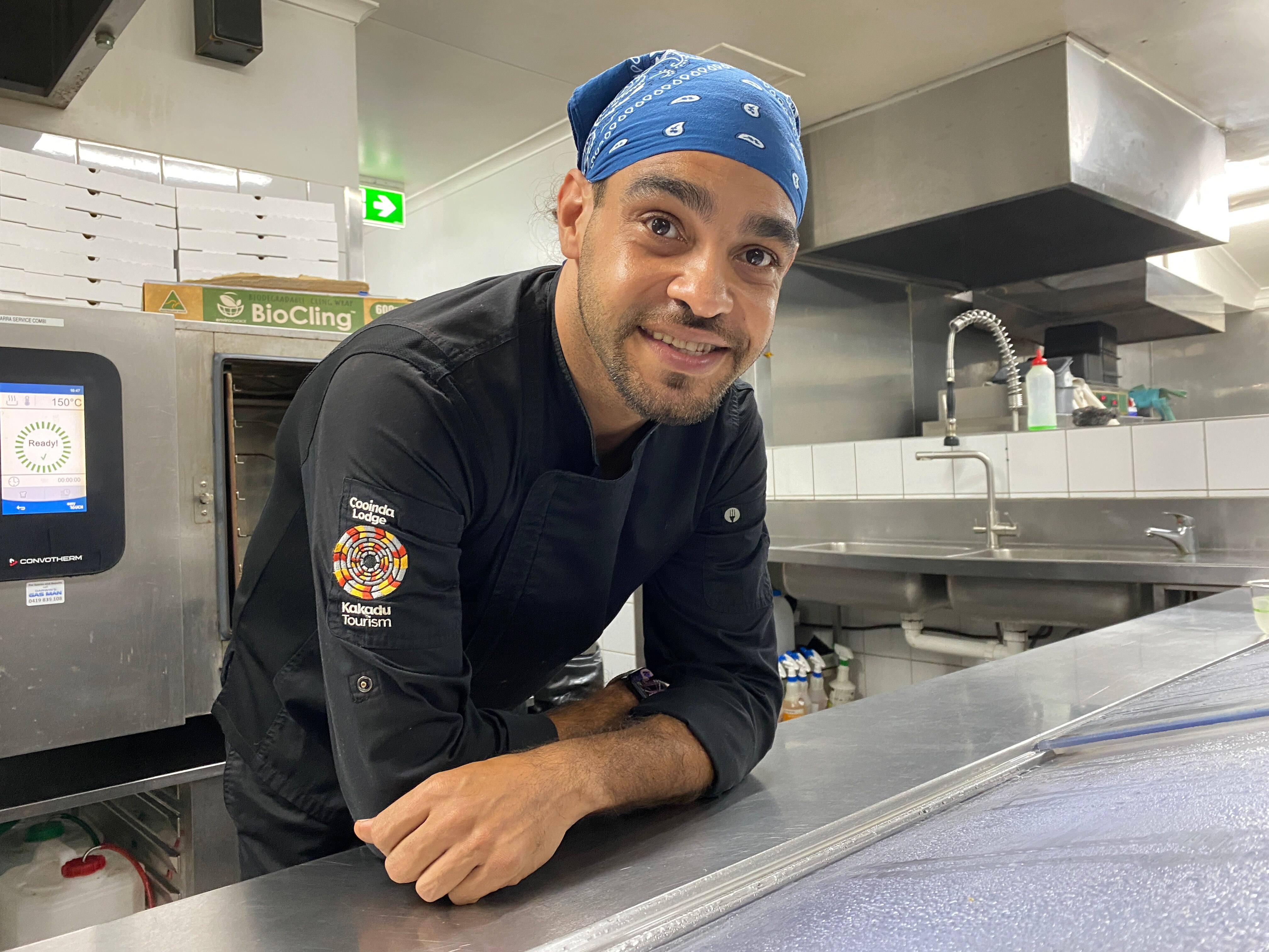 A man leans on a silver counter top in a commercial kitchen wearing black chef's clothes and blue hair cap.