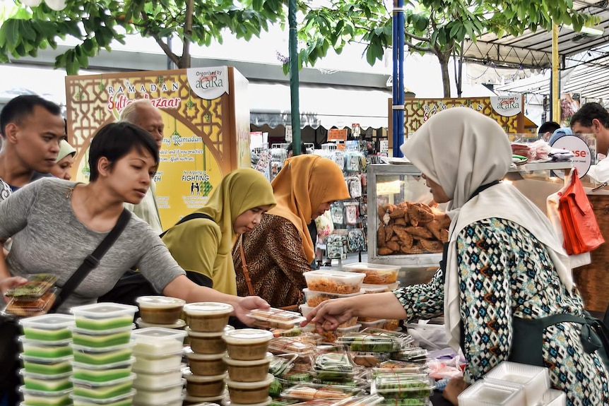 You see a vibrant market stall packed with food items as people are interspersed between the aisles buying goods.