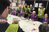 Australian Electoral Commission staff, watched by scrutineers, sit at a large table counting votes.