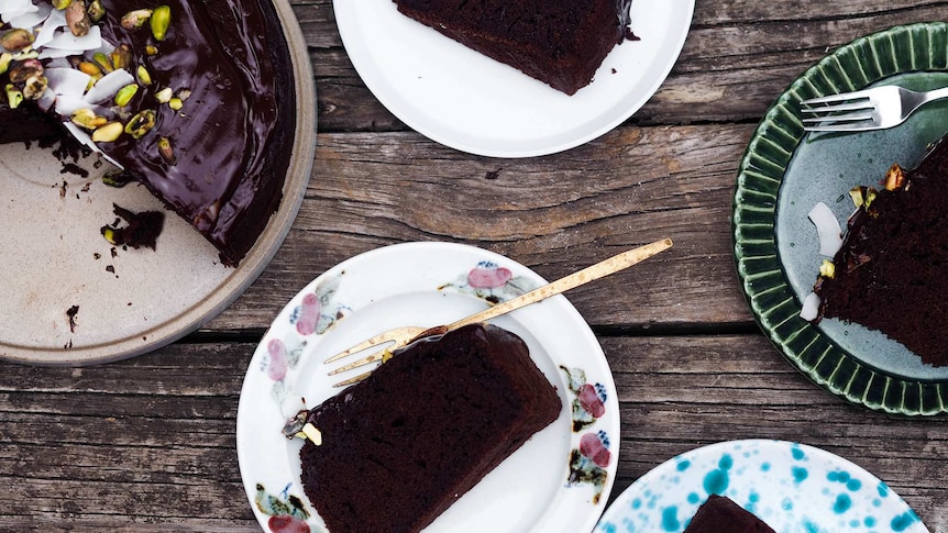 Four slices of chocolate olive oil on plates next to cut cake topped with pistachios and coconut.