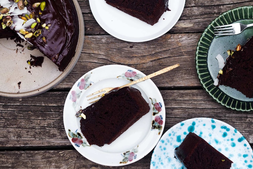 Four slices of chocolate olive oil on plates next to cut cake topped with pistachios and coconut.