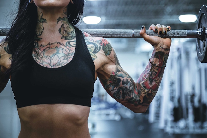 A woman with tattoos in a black sports bra doing weights, her face isn't shown