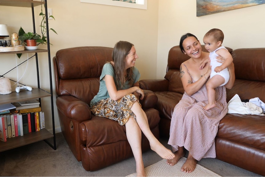 Two women sitting on brown leather lounge chairs, one in a dress holds a baby up, all smiling.