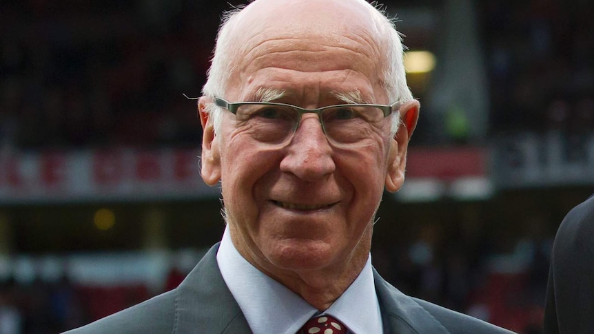 Bobby Charlton smiles at the camera wearing a grey suit with a red and white tie