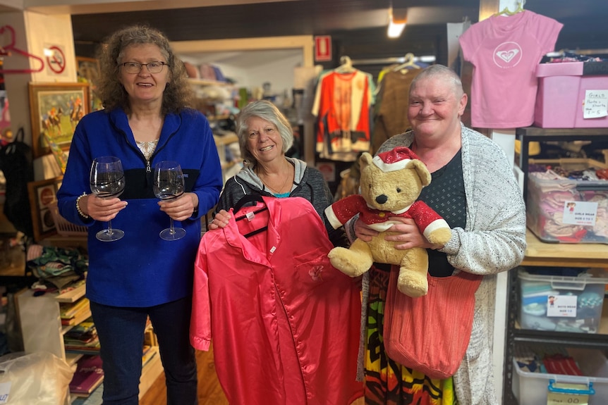 Three women standing holding glasses, a shirt and a teddybear in op shop smiling at camera.