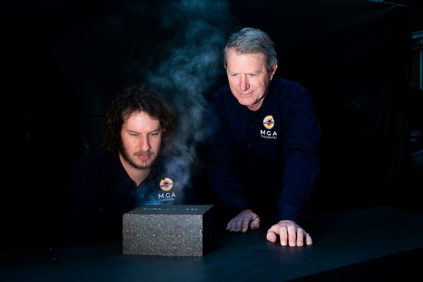 Two men in a studio photograph looking at a large black brick with steam appearing to rise indicating heat