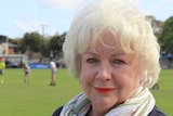 White haired Caucasian woman stands in front of football field where players play.