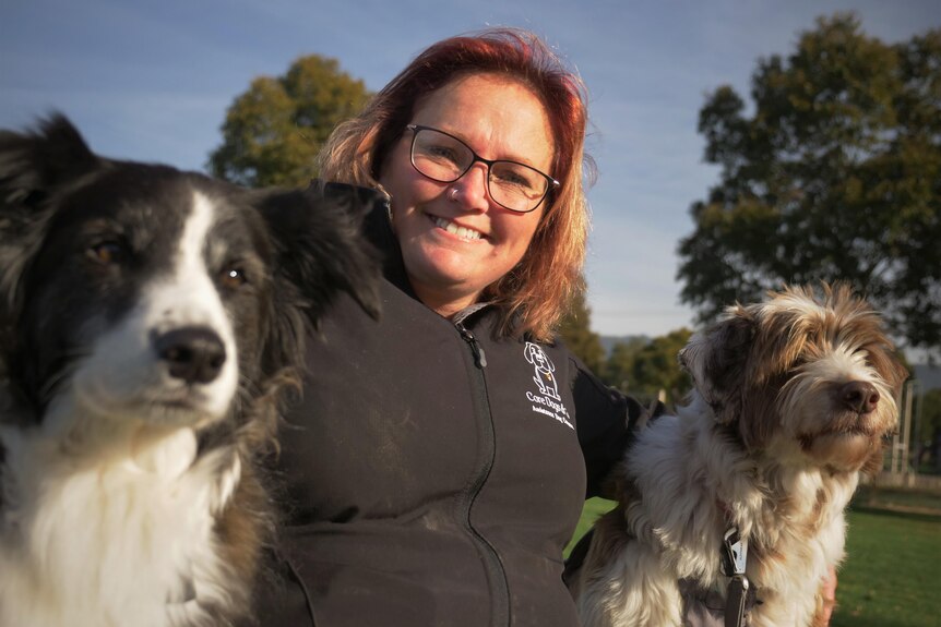 A woman smiling to camera while having her arms over the shoulders of two dogs.