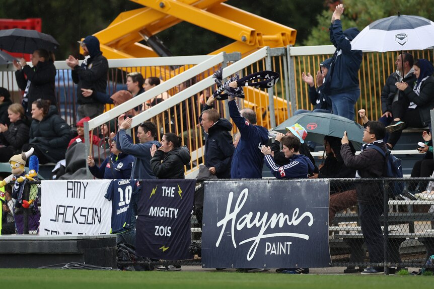Fans wearing rain gear sit and stand on some benches during a sports game