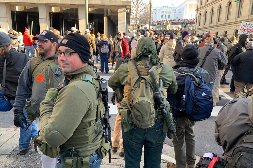 a man wearing a gun on his back looks to the side at the camera as people gather in streets in Richmond, Virginia