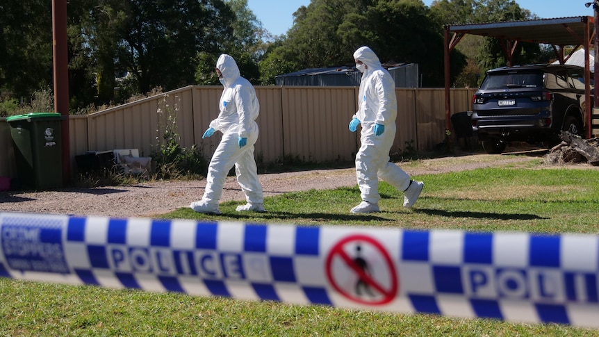 A pair of police officers in hazmat suits walk across the front lawn of a property cordoned off with tape.
