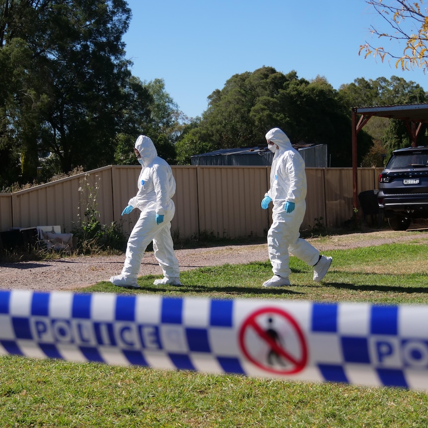 A pair of police officers in hazmat suits walk across the front lawn of a property cordoned off with tape.