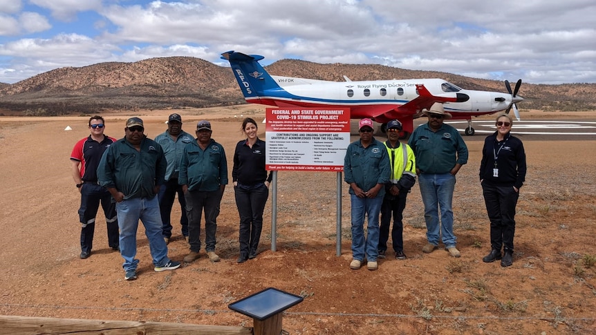 A group of people standing on either side of a sign, with an aircraft on an airstrip in the background.