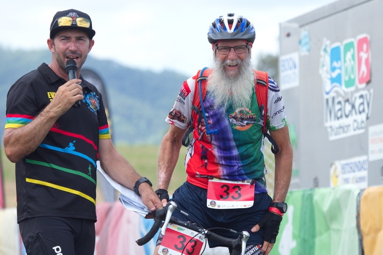 Two men at the finishing line of an event, one on a bike and one with a microphone.