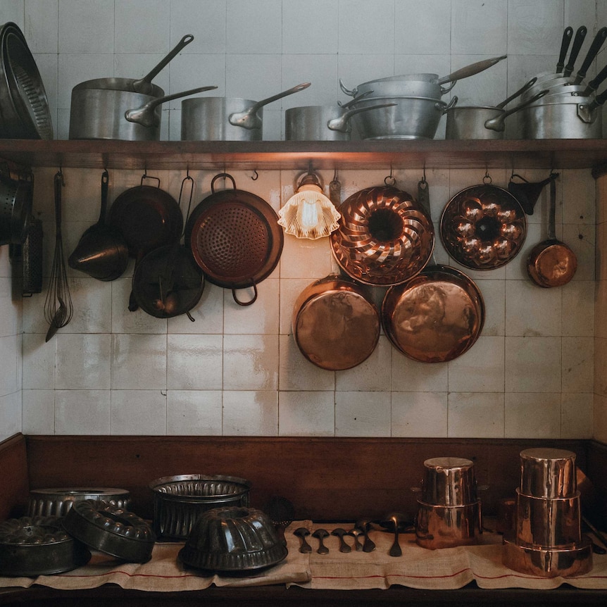 A white-tiled kitchen bench and shelf shows clusters of silver and copper pots and pants.