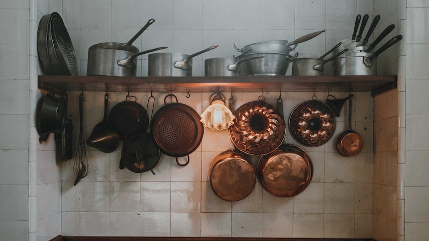 A white-tiled kitchen bench and shelf shows clusters of silver and copper pots and pants.