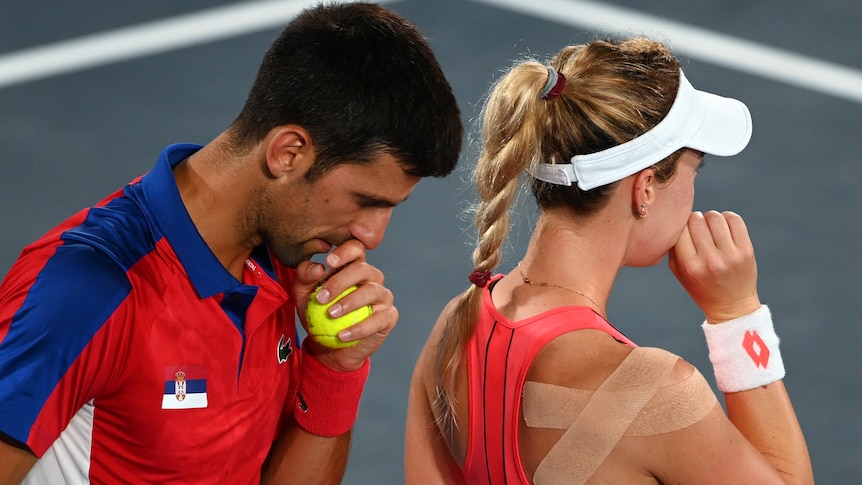 Novak Djokovic holds a tennis ball near mouth as he speaks to mixed doubles partner Nina Stojanovic at the Tokyo Olympic Games.