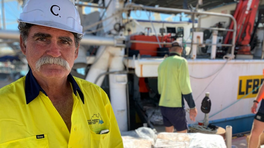 A man in  a hard hat stands next to a fishing trawler.