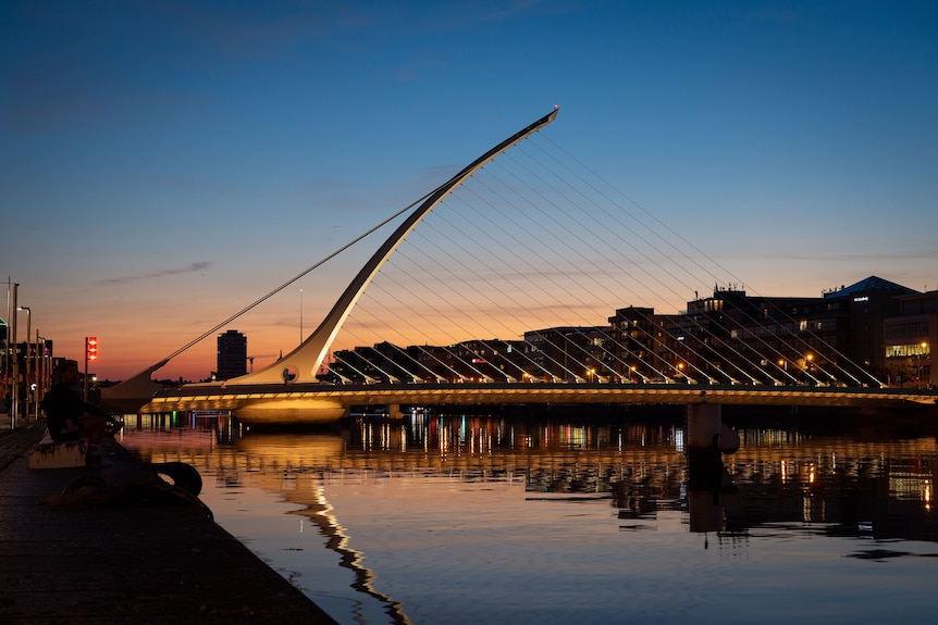 A sunset cityscape showing a harp-shaped bridge spanning a river 