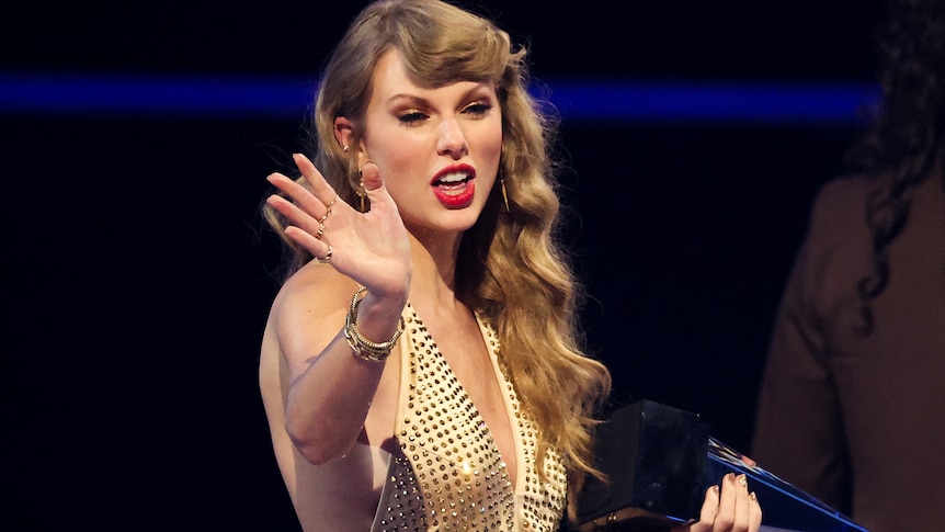 Taylor Swift waving with long blonde hair and red lipstick holding a trophy and wearing a formal dress