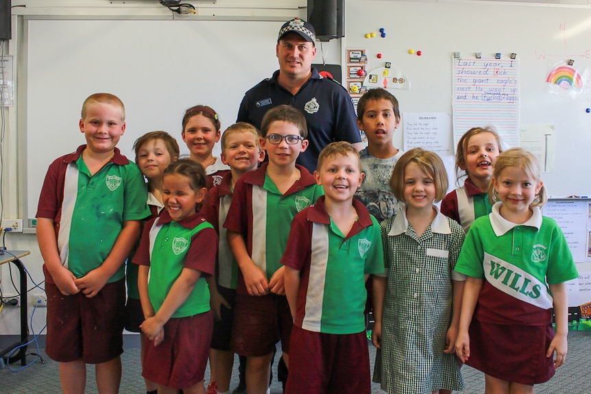 A police officer stands behind young school students wearing green, white and maroon uniforms.