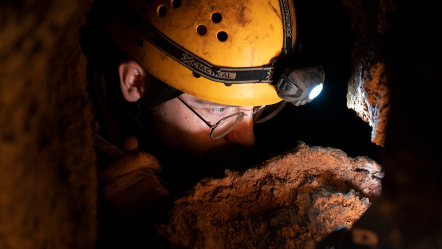 A close up of a man surrounded by rocks. He's wearing a caving helmet with a light. It looks cramped. 