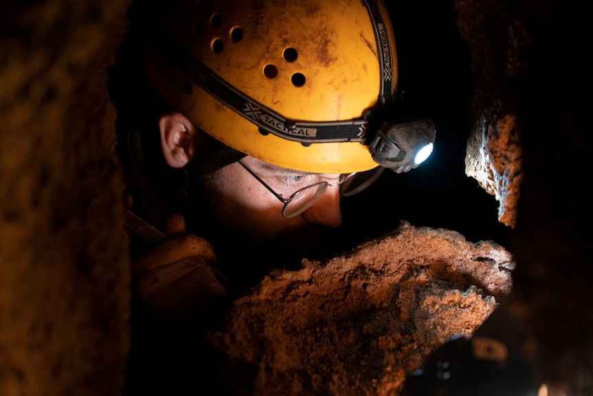 A close up of a man surrounded by rocks. He's wearing a caving helmet with a light. It looks cramped. 
