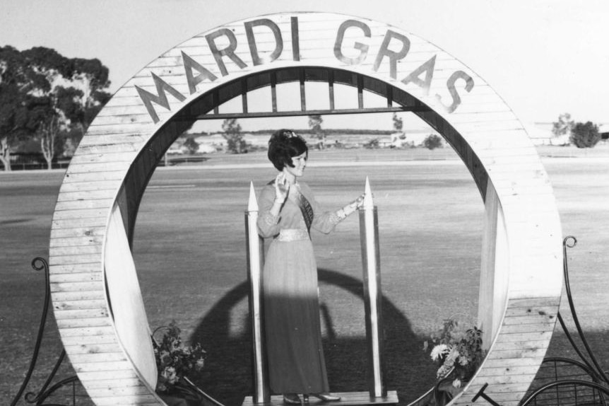 A black and white photo of a woman standing in a round float that has 'Mardi Gras' written on it.