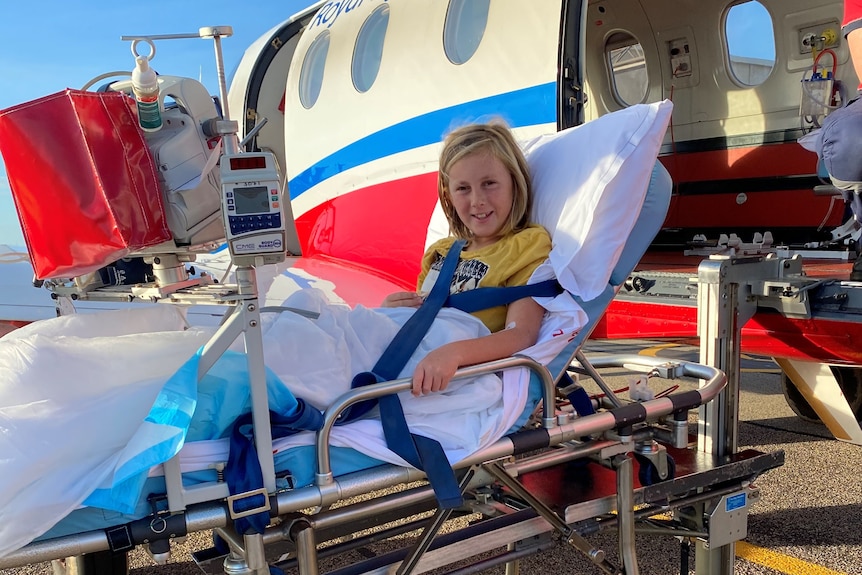 A boy on a medical gurney smiles in front of an RFDS plane on the tarmac