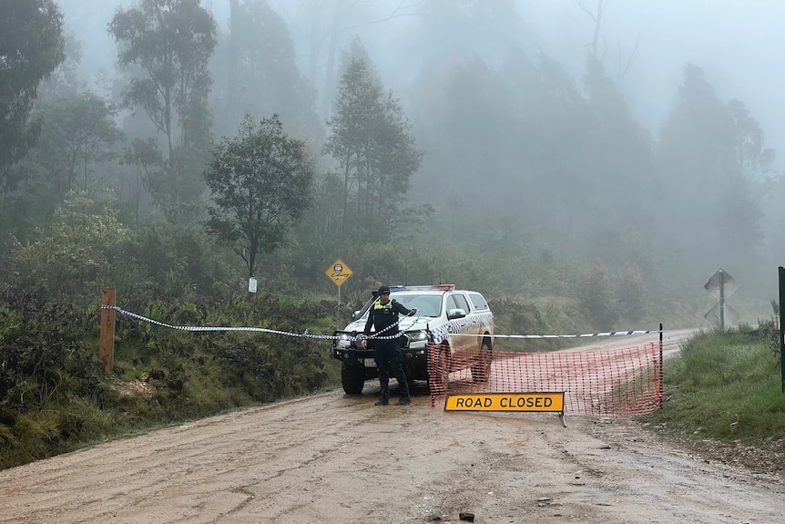 A policeman putting up a police tape to block a road, with a road closed sign, surrounded by trees, mist.