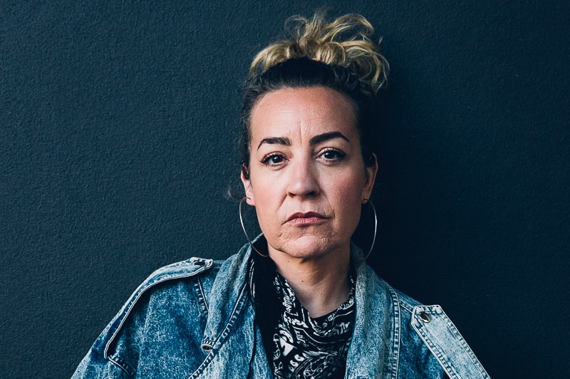 Young woman in denim jacket in front of dark blue wall.