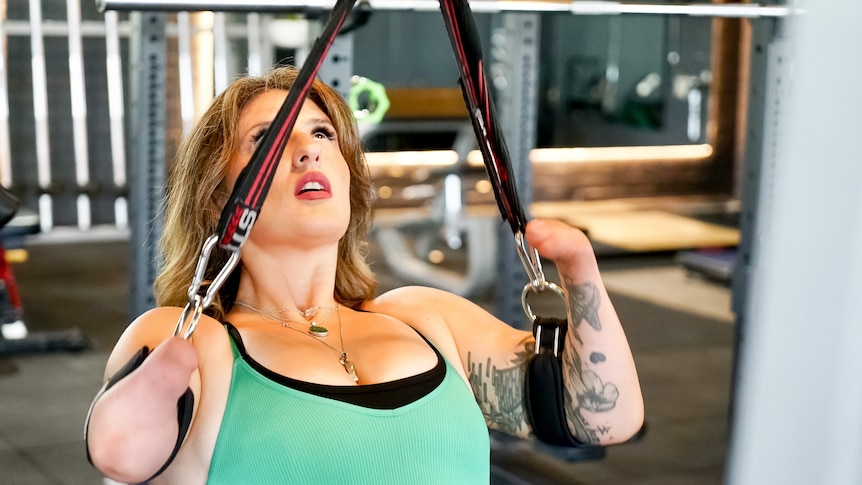 A woman in a green singlet performs pull ups at the gym using two straps. She was born with no hands 