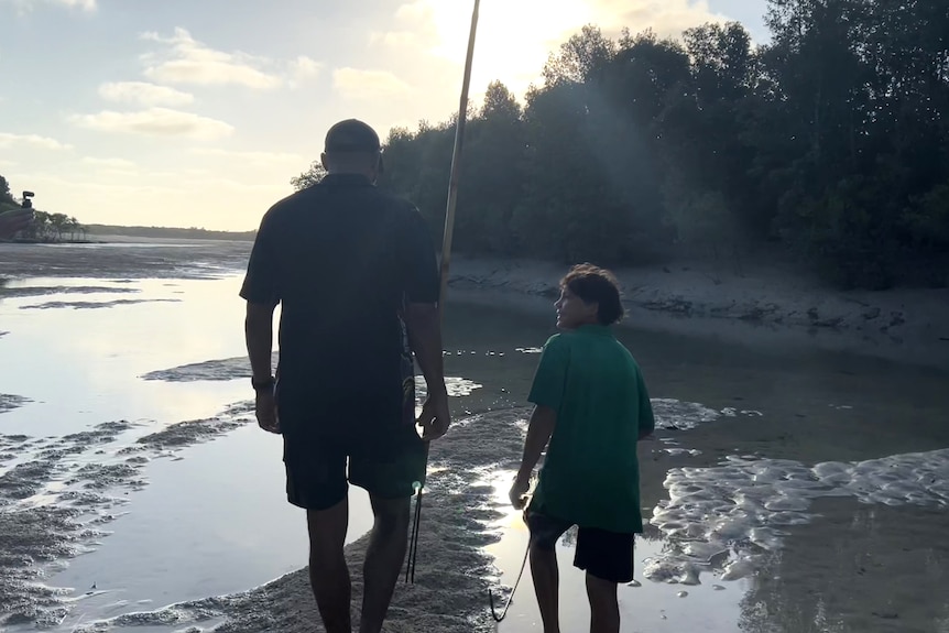 A man walking next to a boy through a sandy creek with mangrove trees around them.