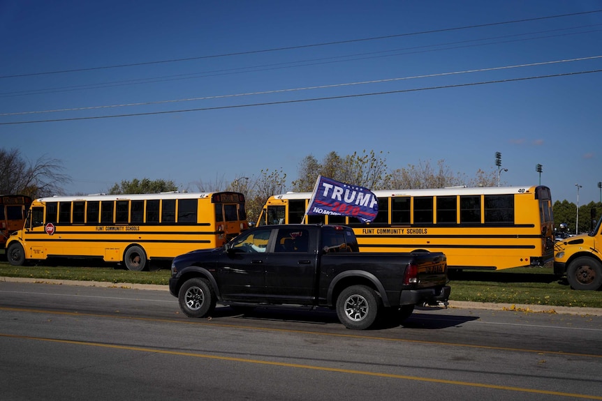 A black truck waving a Trump flag out the window drives passed a parked row of yellow school buses