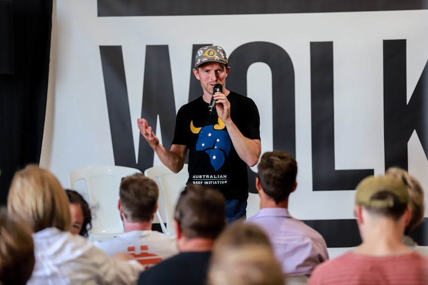 Man in cap and black t-shirt with bull graphic stands on stage speaking to a group of seated people