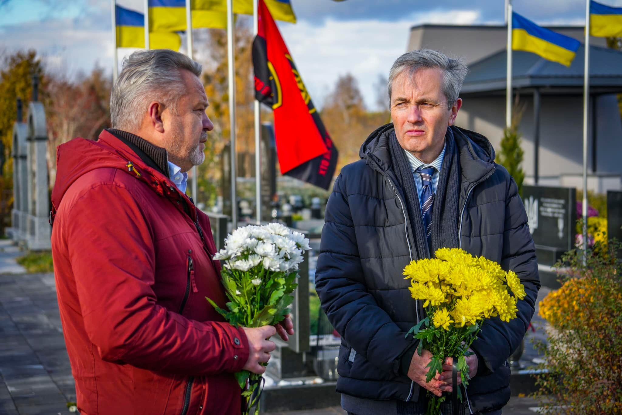 A man in a red jacket holding white flowers speaks to a man in a grey jacket holding yellow flowers