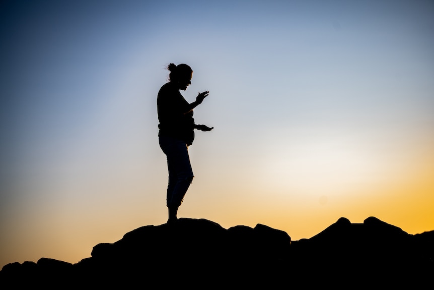 The sillouette of Raelene Cooper, standing on a rock, clapping, as sunrises.