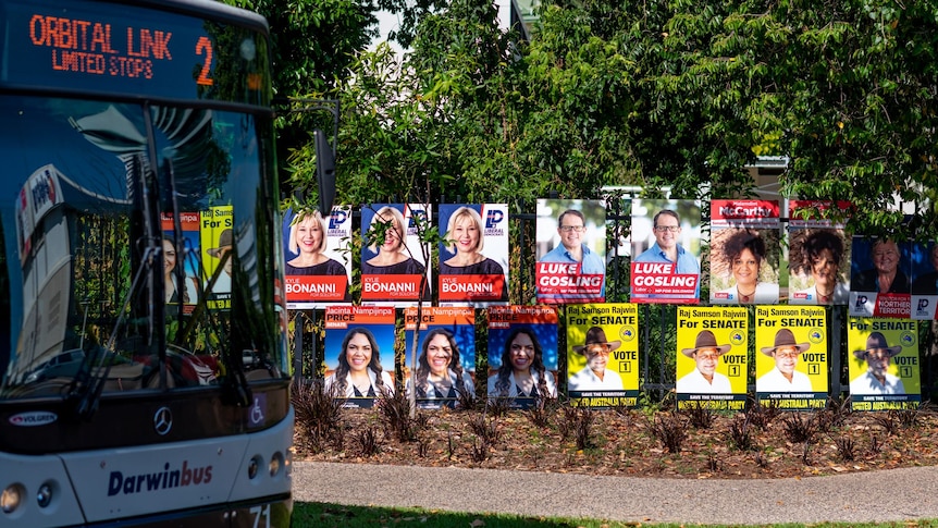A bus driving past a fence covered in posters of federal election candidates.