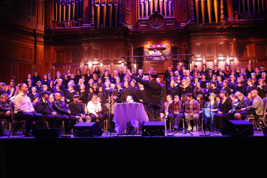 A large choir sits on a stage against a wooden backdrop