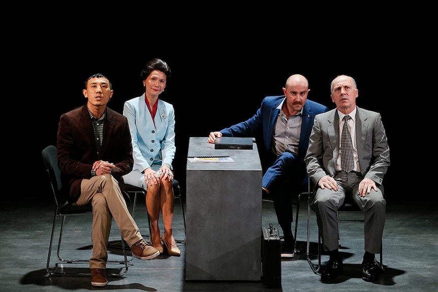 Four people in formal office attire sit in groups of two on either side of slender desk on stage, all face towards audience.