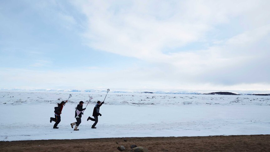 3 people run with lacrosse sticks above their heads in a vast snowy tundra.