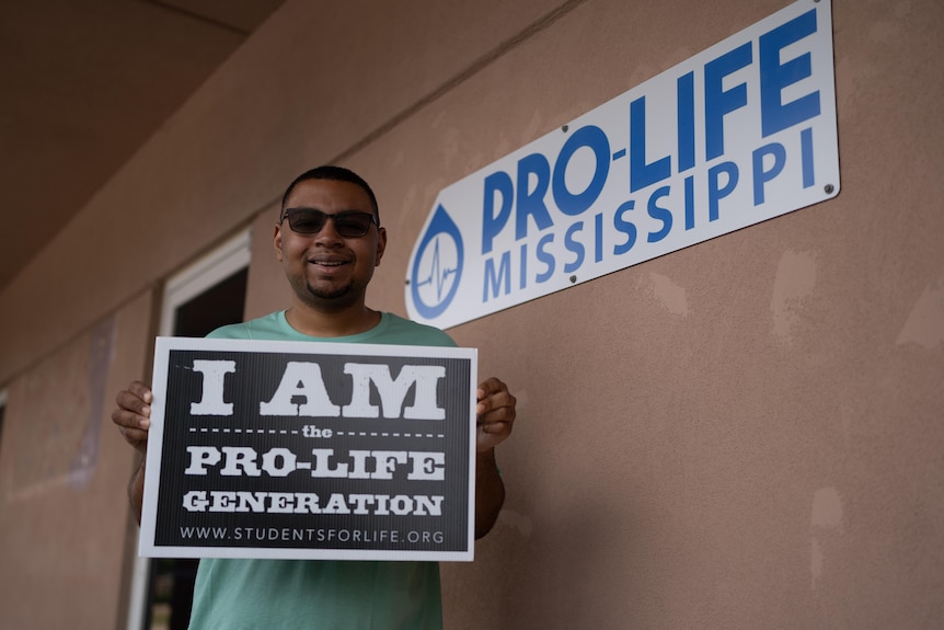 A man stands outside next to a PRO-LIFE MISSISSIPPI sign with a cardboard sign that declares "I am the pro-life generation"