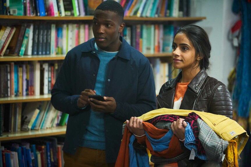 A young black man with a phone and young brown woman holding a stack of clothes, in a library