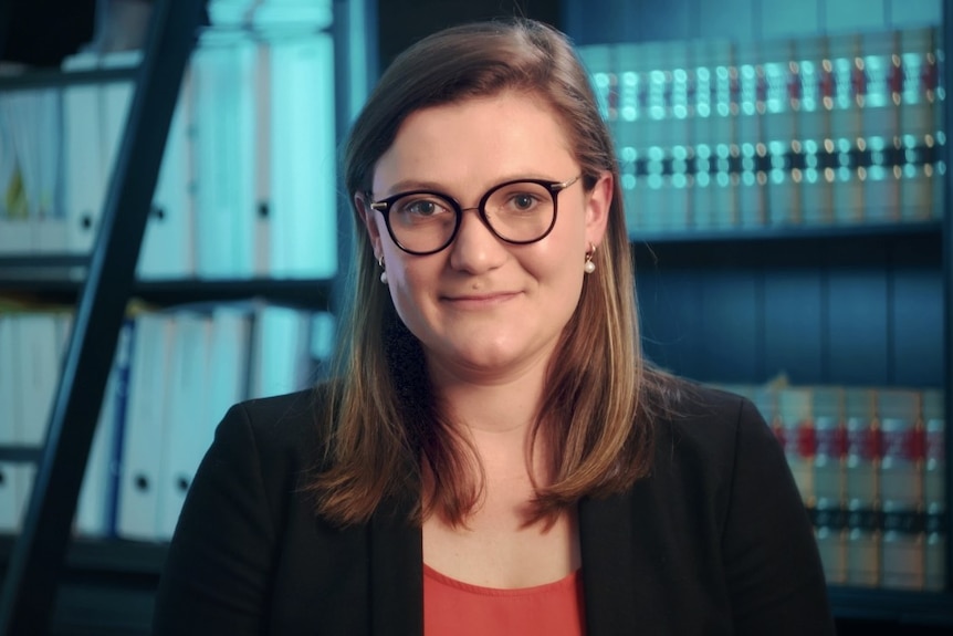 A young woman wearing glasses and a black blazer, with legal books behind her.