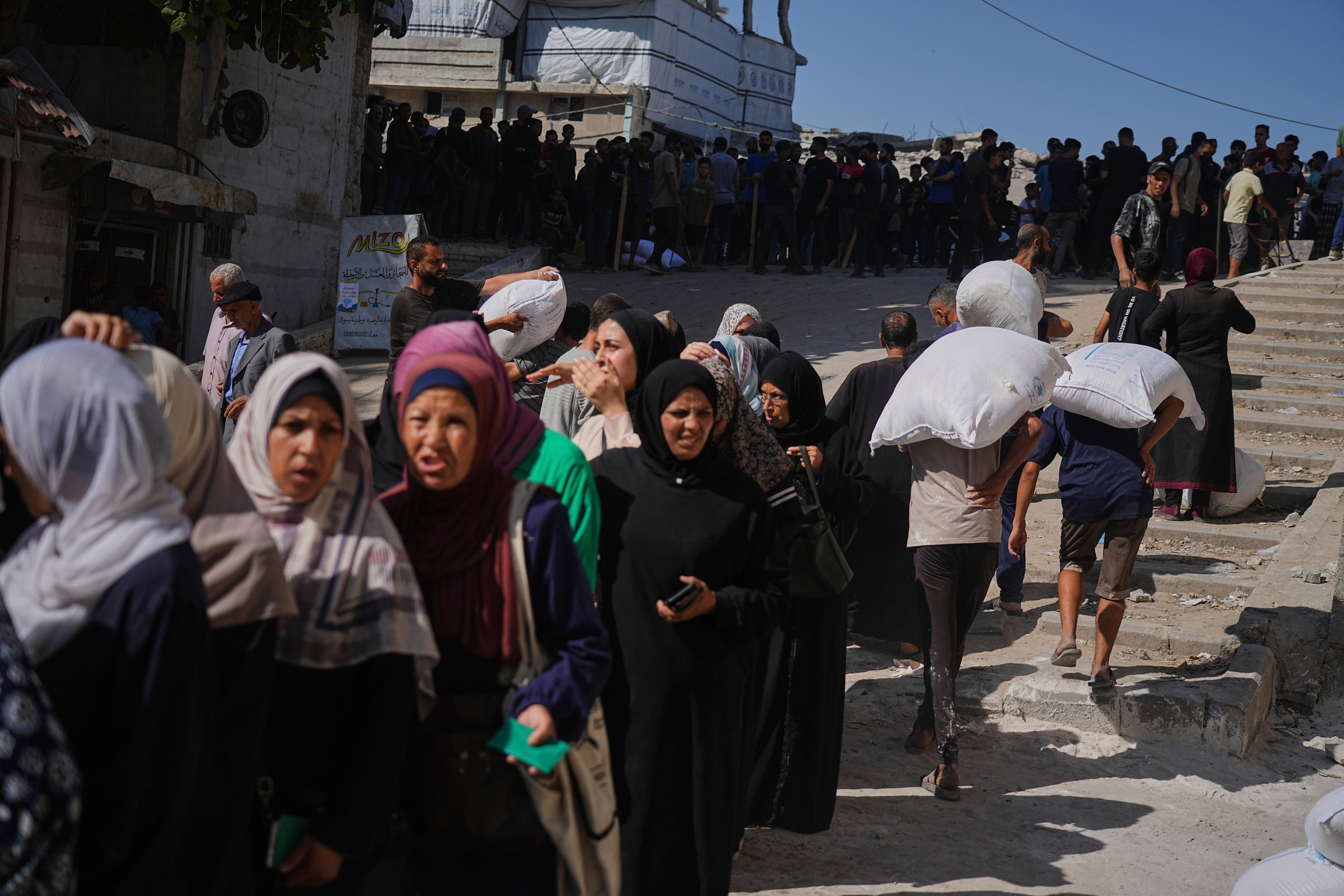 Women wearing black burkhas lined up. In the background some men walk with white bags loaded on their backs