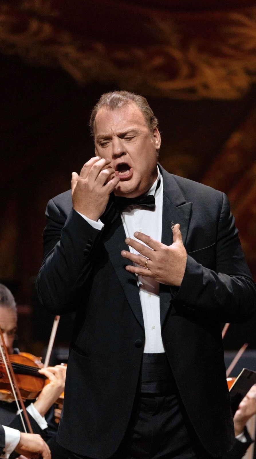 Sir Bryn Terfel performs onstage in a tailcoat, singing with orchestra musicians visible behind him.