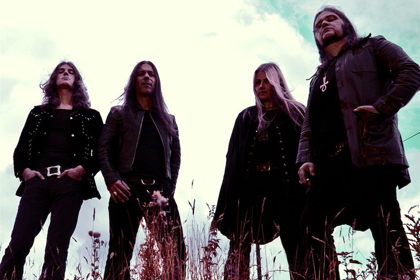 Three men and one women, all with long hair and dressed in dark 70s-era clothing, stand in a field against a moody sky.