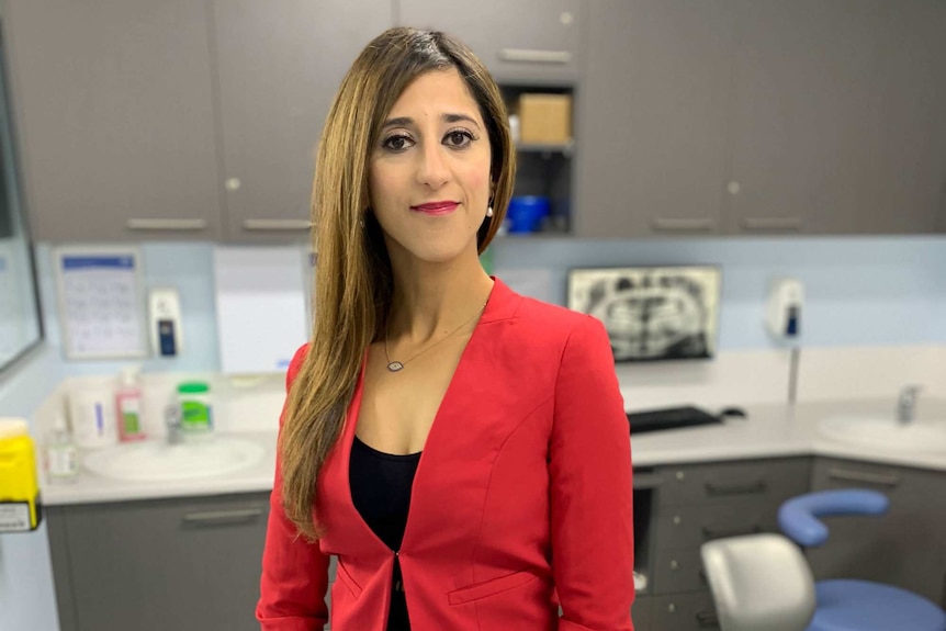 Norah Ayad, wearing a red suit jacket, stands in a dental surgery.