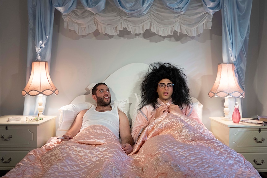 Two men sit in bed in a pale pink and blue satin decorated room, one with comedy expression, the other wearing wig and glasses.